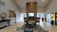 This single-floor space makes an immediate impression with its dramatic vaulted ceilings that amplify the sense of openness and grandeur. The centerpiece of the main lounge area is a striking floor-to-ceiling wood-paneled accent wall, which houses a sleek electric fireplace and a mounted flat-screen television, creating a bold and functional focal point. The seating arrangement is thoughtfully curated, featuring a pair of upholstered wingback chairs flanking the fireplace, complemented by a modern sofa and a geometric-patterned area rug in navy and gold tones that anchors the space. Light-toned ceramic tile flooring runs throughout, contributing to the bright and airy atmosphere. Artistic touches are woven throughout the room, including a sculptural figurine and framed wall art, lending the space a refined and cultured ambiance. Large glass doors and windows at the far end allow natural light to pour in while offering a glimpse of the outdoors. The neutral gray walls serve as a clean backdrop, allowing the wood accents and decor to stand out. Overall, this space balances modern design with a warm, welcoming atmosphere well-suited for gathering and relaxation.