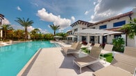This single-story property showcases a striking resort-style outdoor environment centered around a generously sized swimming pool with clear, turquoise water. The pool deck is finished in large-format light-toned porcelain or stone tiles, providing a clean, polished surface that reflects the abundant natural sunlight. Arranged along the deck are modern white-framed lounge chairs with tan mesh seating, complemented by bold black-and-white striped market umbrellas that add a sophisticated, contemporary touch.

The surrounding building exhibits a Mediterranean or Spanish Colonial architectural style, characterized by a terracotta-tiled roofline, exposed decorative wood beam detailing along the eaves, and a crisp white stucco exterior. Large windows and glass doors open directly to the pool area, creating a seamless connection between the interior and outdoor living spaces.

Lush tropical landscaping, including mature palm trees and vibrant foliage, frames the perimeter, enhancing the resort ambiance. The combination of the bright open sky, reflective pool water, and carefully curated outdoor furnishings creates an inviting, upscale atmosphere well-suited for relaxation and leisure.