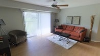 This single-floor residence offers a welcoming and functional layout centered around a bright, open living area. The space is finished with warm hardwood-style flooring that extends throughout, lending a clean and cohesive look to the interior.

The living room is a standout feature, bathed in natural light streaming through expansive floor-to-ceiling sliding glass doors dressed with vertical blinds. These doors provide a seamless connection to the outdoors while allowing full control over light and privacy. A ceiling fan with an integrated light fixture is centrally positioned overhead, promoting airflow and comfort across the space.

The room comfortably accommodates a tufted sofa, accent seating, and a pair of sleek ottomans arranged over a plush area rug, illustrating the generous proportions of the living area. Wall art adds a decorative touch, complementing the neutral gray tones of the walls.

Overall, the space presents a functional yet stylish single-floor layout with an emphasis on natural light, open living, and practical design—making it a comfortable and inviting environment for everyday living.