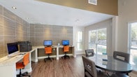 This business center is a thoughtfully designed, single-floor space that blends functionality with a contemporary aesthetic. The layout is divided into distinct zones, allowing for focused work and relaxed collaboration simultaneously. Warm hardwood-style flooring runs throughout, anchoring the space with a cohesive, polished look.

The workstation area is a standout feature, set against a full-height textured tile accent wall in muted earth tones. A built-in granite-topped desk accommodates multiple computer workstations, complemented by vibrant orange ergonomic chairs that add a modern pop of color. A wall-mounted flat-screen television and additional floating desk provide supplementary workspace nearby.

The lounge and meeting area offers a round glass-top table with upholstered chairs alongside comfortable black leather armchairs paired with side tables, creating an inviting setting for informal gatherings or individual use. A ceiling fan overhead enhances comfort throughout the open area.

Natural light is abundant, with large windows and a sliding glass door framing views of the outdoor pool and landscaped surroundings. An additional windowed seating nook along the perimeter further maximizes these serene outdoor views, giving the space an open, airy ambiance well-suited to a productive yet relaxed environment.
