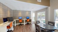 This business center is a thoughtfully designed, single-floor space that blends functionality with a contemporary aesthetic. The layout is divided into distinct zones, allowing for focused work and relaxed collaboration simultaneously. Warm hardwood-style flooring runs throughout, anchoring the space with a cohesive, polished look.

The workstation area is a standout feature, set against a full-height textured tile accent wall in muted earth tones. A built-in granite-topped desk accommodates multiple computer workstations, complemented by vibrant orange ergonomic chairs that add a modern pop of color. A wall-mounted flat-screen television and additional floating desk provide supplementary workspace nearby.

The lounge and meeting area offers a round glass-top table with upholstered chairs alongside comfortable black leather armchairs paired with side tables, creating an inviting setting for informal gatherings or individual use. A ceiling fan overhead enhances comfort throughout the open area.

Natural light is abundant, with large windows and a sliding glass door framing views of the outdoor pool and landscaped surroundings. An additional windowed seating nook along the perimeter further maximizes these serene outdoor views, giving the space an open, airy ambiance well-suited to a productive yet relaxed environment.