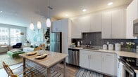 This contemporary single-floor residence is thoughtfully designed with modern finishes and an open, functional layout. The kitchen serves as a central highlight, featuring white shaker cabinetry extending to the ceiling, granite countertops, stainless steel appliances including a full-size refrigerator, dishwasher, microwave, and range, along with a grey subway tile backsplash and recessed lighting. Pendant lights illuminate a freestanding island with seating, making it ideal for casual dining. Wood-style plank flooring flows seamlessly from the kitchen into the living area, which is bathed in natural light through large windows overlooking the surrounding community. The living space offers a generous, flexible layout suited for both relaxing and entertaining. The bedroom is a serene retreat, featuring carpet flooring, a ceiling fan, and wide windows that invite abundant natural light. It provides convenient access to a full bathroom appointed with granite vanity countertops, white cabinetry, and a full tub-shower combination. Clean lines, neutral tones, and consistent modern finishes throughout create a cohesive and welcoming ambiance across the entire residence.