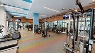 Located in Miramar, FL, this modern fitness center offers a generously sized, single-floor layout designed to accommodate a comprehensive range of workout needs. The space features a bright, open floor plan with large floor-to-ceiling windows and glass doors that flood the interior with natural light and provide views of lush outdoor greenery, creating an energizing atmosphere for exercise.

The facility is divided into clearly defined workout zones. The cardio area is lined with treadmills, ellipticals, and stair climbers, all positioned to take advantage of the natural light and outdoor views. The strength training section is well-appointed with a multi-station cable machine, free weights including a full rack of dumbbells, a power rack, medicine balls, a punching bag, TRX suspension trainers, and adjustable benches — catering to both functional and traditional training styles.

Architecturally, the space features warm wood-paneled accent walls, vibrant teal structural columns, and linear recessed lighting that runs along a crisp white ceiling with colorful geometric accents. Full-length mirrors line several walls, enhancing the sense of openness and allowing for form checks during exercise. Colorful, patterned carpet tiles add a dynamic visual energy to the floor throughout. Wall-mounted flat-screen televisions are positioned for easy viewing from multiple workout stations, completing this thoughtfully designed fitness facility.