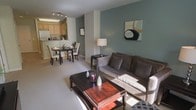 This single-floor residence showcases an open-concept floor plan that integrates the living, dining, and kitchen areas into one cohesive and functional space. The living area is anchored by a plush gray sofa paired with a rich, dark-toned wooden coffee table, creating an inviting focal point. A muted teal accent wall adds a pop of color and depth to the space, complemented by neutral gray walls and light-toned carpeted flooring throughout.

Adjacent to the living area, the dining space accommodates a dark wood table surrounded by upholstered chairs, ideal for everyday meals or entertaining. A delicate floral centerpiece adds a touch of warmth. Natural light streams into the room, enhancing the open and airy feel of the layout.

The kitchen, visible from both the living and dining areas, features light wood cabinetry and white appliances, offering a clean and functional workspace. Overhead track lighting ensures the kitchen is well-lit and practical. The seamless flow between all three zones makes this space efficient and well-suited for comfortable everyday living.