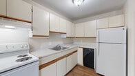 This single-floor residential unit offers a practical and comfortable layout designed for everyday living. The kitchen is well-equipped with white cabinetry accented by warm wood trim, light-toned laminate countertops, a stainless steel sink, a white electric range, a black dishwasher, and a full-size refrigerator. Rich hardwood-style flooring runs throughout the kitchen, adding warmth to the space.

The living area is generously sized, featuring plush wall-to-wall carpeting, a ceiling fan with integrated lighting, baseboard heating, and a wall-mounted air conditioning unit for year-round comfort. Large vertical blinds cover a sizable window, allowing for adjustable natural light. The open feel of the living space is enhanced by neutral-toned walls that create a clean, bright atmosphere.

The bedrooms continue the carpeted flooring and neutral color palette, each benefiting from natural light through vertical-blind-covered windows and baseboard heating. Closet space is present, providing practical storage. The bathroom features a full tub and shower combination with clean white tile surround, offering a simple and functional design. Overall, this unit presents a straightforward, move-in-ready layout with consistent finishes throughout.