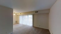 This single-story space features an open living area designed to maximize comfort and functionality. The room is finished with wall-to-wall carpeting in a neutral tone, providing a soft and cohesive foundation throughout. A large sliding glass door spans a significant portion of one wall, fitted with vertical blinds that allow flexible control of natural light while offering access to an outdoor area. The generous window presence ensures the interior remains bright and airy. A ceiling fan is centrally positioned, contributing to efficient air circulation year-round. HVAC vents are strategically placed along the ceiling and walls, indicating a comprehensive climate control system. The walls are finished in a light, neutral palette that enhances the sense of openness. Baseboard details and clean wall lines give the space a simple, unfussy character. The layout is open and unobstructed, offering versatile possibilities for furniture arrangement and use of the area. Overall, the space presents a straightforward, well-lit environment with practical features suited for everyday living.