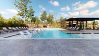 Located at 3000 Fast Ln in Fort Mill, SC, this outdoor amenity space is designed to deliver a resort-style experience within a contemporary residential setting. The centerpiece is a generously sized swimming pool with crystal-clear water, flanked by rows of lounge chairs positioned along a smooth concrete pool deck. Mature trees line the perimeter, providing natural shade and a sense of seclusion beneath open skies.

Adjacent to the pool, a spacious paver patio offers a versatile social hub, furnished with modern seating arrangements including chairs and tables ideal for outdoor dining or casual gatherings. Overhead string lights add ambiance, creating an inviting atmosphere during evening hours. A pergola-style structure with additional lounge seating anchors one end of the pool area, extending the space for relaxation and shade.

The building entry features a striking façade with soaring floor-to-ceiling glass doors framed by substantial architectural columns and a mix of stone and brick cladding. Carefully maintained turf panels and manicured landscaping beds with ornamental plantings add greenery and visual interest throughout the courtyard. The overall design balances modern architectural lines with natural elements, creating a well-appointed outdoor environment suited for both active use and leisurely enjoyment.