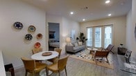 This single-floor space features an open-plan layout that seamlessly connects the dining and living areas, creating a cohesive and functional flow. The dining area is anchored by a round marble-top table surrounded by warm tan upholstered chairs, complemented by decorative woven wall plates that add a touch of cultural character. The living area is furnished with a gray sofa and coordinating accent chairs arranged on a patterned area rug, centered around a round coffee table. Large windows with blinds line one wall, allowing generous natural light to fill the space while offering views to the exterior. Recessed lighting throughout provides consistent illumination. Light wood-toned flooring runs across the entire living space, lending warmth and continuity. A doorway connects the open living area to a separate bedroom, visible in the background with a neatly made bed and darker toned furnishings. The neutral wall palette and clean architectural lines give the space a modern, well-organized feel suited for comfortable everyday living.