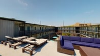 This amenity space comprises two distinct areas designed to support an active lifestyle. The sun deck is an open-air rooftop terrace offering expansive views of the surrounding community. The deck features wicker chaise lounges and a sectional sofa with navy blue cushions, providing comfortable spaces for relaxing or socializing under open skies. Metal railings line the perimeter, and the outlook includes neighboring buildings and lush greenery below.

The fitness center is a generously sized, well-lit space with high ceilings adorned with crown molding and ceiling fans. Large windows along multiple walls invite abundant natural light and views of the outdoors, creating an airy workout environment. The flooring features a herringbone-patterned carpet in blue and grey tones throughout. A floor-to-ceiling mirror along one wall visually expands the space.

Equipment is comprehensive, including a multi-station cable and power rack system, a full dumbbell rack, medicine ball storage, battle ropes, a Precor rowing machine, a WaterRower, a cable crossover machine, a squat rack with bench, and multiple treadmills with an integrated wall-mounted TV. The combination of strength and cardio equipment makes this fitness center suitable for a wide range of training needs.