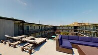 This amenity space comprises two distinct areas designed to support an active lifestyle. The sun deck is an open-air rooftop terrace offering expansive views of the surrounding community. The deck features wicker chaise lounges and a sectional sofa with navy blue cushions, providing comfortable spaces for relaxing or socializing under open skies. Metal railings line the perimeter, and the outlook includes neighboring buildings and lush greenery below.

The fitness center is a generously sized, well-lit space with high ceilings adorned with crown molding and ceiling fans. Large windows along multiple walls invite abundant natural light and views of the outdoors, creating an airy workout environment. The flooring features a herringbone-patterned carpet in blue and grey tones throughout. A floor-to-ceiling mirror along one wall visually expands the space.

Equipment is comprehensive, including a multi-station cable and power rack system, a full dumbbell rack, medicine ball storage, battle ropes, a Precor rowing machine, a WaterRower, a cable crossover machine, a squat rack with bench, and multiple treadmills with an integrated wall-mounted TV. The combination of strength and cardio equipment makes this fitness center suitable for a wide range of training needs.