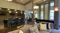Located at 8300 Western Blvd in Raleigh, NC, this single-floor space is designed with an open-concept layout that seamlessly connects multiple functional zones. The interior is anchored by striking stacked-stone columns that add a rustic yet refined architectural element, complementing the warm, neutral color palette throughout.

The lounge area features plush, generously sized seating adorned with decorative pillows in navy, yellow, and patterned accents, centered around a wooden coffee table. Overhead, a combination of pendant lights and a chandelier provides layered, ambient illumination that enhances the inviting atmosphere.

Adjacent to the lounge, a bar-height dining area is outfitted with sleek charcoal chairs and a wall-mounted television, making it well-suited for social gatherings. A billiards table positioned near tall, draped windows introduces a recreational element, while natural light filters in through the patterned curtains, brightening the space.

Soaring ceilings with coffered detailing and recessed lighting add to the sense of grandeur, while the dark flooring provides a sophisticated contrast to the lighter walls and furnishings. Overall, the space exudes a polished, welcoming ambiance ideal for community or social use.