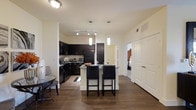 This single-story residence features a thoughtfully designed open-concept layout that seamlessly connects the living, dining, and kitchen areas. Rich hardwood flooring flows throughout the main living space, grounding the interior with warmth and continuity.

The kitchen is a standout feature, outfitted with dark espresso cabinetry that extends to the upper walls, providing generous storage. A light-toned countertop island serves as the centerpiece, offering additional prep space and casual seating for two with sleek black bar stools. Pendant lighting hangs above the island, complementing the track lighting overhead to create a layered and functional lighting scheme.

The open layout allows for a natural transition into the living area, where a glass-top accent table adds a contemporary touch. A corridor leads directly to the bedroom, which is visible through a doorway, hinting at a private and well-separated sleeping area. A large paneled closet door nearby provides practical storage within the main living space.

The overall ambiance is modern and cohesive, with a neutral wall palette that allows the dark cabinetry and warm flooring to serve as the primary design elements.