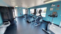 This fitness center is a single-floor facility designed to support a well-rounded workout experience. The interior is defined by its bold teal-painted walls, which lend the space a modern and energetic atmosphere, complemented by dark rubberized flooring that provides both durability and safety during exercise.

The space is outfitted with a variety of cardio equipment, including treadmills, an elliptical trainer, and what appears to be a rowing machine, offering users multiple options for aerobic training. A multi-station cable and weight machine anchors the strength training area, accompanied by a dedicated bench, making it suitable for a range of resistance exercises.

Natural light flows generously into the room through a series of large windows along one wall, brightening the space and providing views of an outdoor sports court and surrounding greenery. Overhead fluorescent lighting supplements the natural light, ensuring the room remains well-lit at all times.

Practical safety features are also present, including a mounted fire extinguisher and clearly marked emergency exit. The layout is open and thoughtfully arranged, allowing for comfortable movement between equipment. Overall, this fitness center delivers a functional and inviting environment for residents or guests seeking convenient access to exercise facilities.