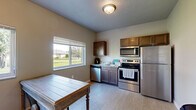 Located at 218 19th St SE in Watertown, South Dakota, this single-floor space offers a well-organized and functional layout across five distinct areas: a kitchen, dining room, office, bathroom, and hallway. The kitchen is a standout feature, equipped with a full suite of stainless steel appliances including a refrigerator, electric range, built-in microwave, and dishwasher. Dark-toned wood cabinetry provides ample storage, while a window with horizontal blinds allows natural light to filter in with views of a green outdoor landscape. Light wood-style flooring runs throughout the main area, lending a clean, cohesive aesthetic. The dining room accommodates a round wooden table with padded chairs, creating an intimate setting for meals. A wall-mounted HVAC unit provides climate control in this area. The office is carpeted and features neutral gray walls, offering a quiet, focused workspace. The bathroom includes a walk-in shower stall with a clean white surround and chrome fixtures. Neutral-toned walls throughout the space contribute to a calm, understated ambiance. The layout makes efficient use of every square foot, making this a practical and well-appointed space suited for day-to-day functional needs.