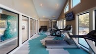 Located at 8625 Winter Oaks Lane in Charlotte, NC, this well-appointed fitness center is designed to deliver a bright, motivating workout environment. The space is defined by its striking vaulted ceilings, which create a sense of openness and airiness throughout. A bold teal and neutral geometric-patterned carpet adds a dynamic visual element underfoot, complementing the warm taupe walls and clean white trim.

Large floor-to-ceiling glass sliding doors and expansive windows line the walls, flooding the interior with natural light and offering views of the surrounding outdoor area. Clerestory windows near the roofline further enhance the luminous atmosphere.

The fitness center is comprehensively equipped for a full-body workout. Cardio options include multiple treadmills and an elliptical machine, while strength training is supported by a multi-station cable machine and a well-stocked dumbbell rack. A wall-mounted flat-screen television is positioned for easy viewing during workouts. Recessed lighting provides consistent illumination throughout the space.

A glass-panel interior door leads to an adjacent room, and a wall-mounted unit provides climate control for year-round comfort. The overall design balances functionality with an inviting, modern aesthetic, making this fitness center a standout amenity for residents of the Winter Oaks Lane community in Charlotte.