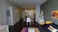 This well-appointed single-floor residence offers a cohesive open-concept design that seamlessly connects the living, dining, and kitchen areas into one fluid, functional space. Dark hardwood floors run throughout, providing a rich, grounding contrast to the bright white walls and ceilings. Recessed lighting illuminates the interior evenly, while a pendant light above the kitchen area adds a focused decorative touch.

The kitchen is positioned toward the rear of the open layout and features stainless steel appliances, including a full-size refrigerator, along with dark cabinetry that adds depth and contrast. Ample counter space and an efficient layout make it both practical and visually appealing.

Adjacent to the kitchen, the dining area is furnished with a white dining table and matching chairs, creating a clean, contemporary look. A colorful area rug anchors the space and adds a vibrant pop of color.

The living area, positioned at the front, is accented by a bold accent wall in deep gray, lending the space a modern, sophisticated character. Decorative accessories and soft furnishings introduce personality throughout. A hallway visible to one side suggests additional private spaces beyond. Overall, the layout is efficient yet thoughtfully designed for comfortable everyday living.