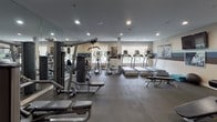 The Bridgewater Fitness Center at 100 Bellis Ct in Bridgewater, NJ is a thoughtfully designed, single-floor facility built to support a comprehensive workout experience. The space is finished with durable dark flooring throughout, recessed lighting, and a distinctive tri-color accent stripe — in shades of light blue, white, and dark brown — that runs along the walls, giving the gym a cohesive and energetic aesthetic. Large mirrors line key sections of the walls, enhancing the sense of openness and allowing members to monitor their form during exercises.

The cardio area is well-stocked with multiple treadmills, elliptical trainers, and stationary bikes, all positioned near windows that allow natural light to filter in and offer views of the surrounding greenery. Strength training is equally well-represented, featuring a multi-station cable and weight machine system, a dedicated free-weight zone with a full dumbbell rack, adjustable benches, and a stability ball. Motivational black-and-white fitness photography is displayed in framed artwork across the walls, contributing to an inspiring atmosphere.

Additional amenities include a wall-mounted flat-screen television, a towel/accessory station, and organized storage cubbies. The facility is designed for functionality and comfort, making it suitable for a wide range of fitness routines.