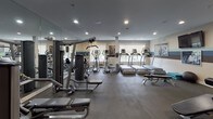 The Bridgewater Fitness Center at 100 Bellis Ct in Bridgewater, NJ is a thoughtfully designed, single-floor facility built to support a comprehensive workout experience. The space is finished with durable dark flooring throughout, recessed lighting, and a distinctive tri-color accent stripe — in shades of light blue, white, and dark brown — that runs along the walls, giving the gym a cohesive and energetic aesthetic. Large mirrors line key sections of the walls, enhancing the sense of openness and allowing members to monitor their form during exercises.

The cardio area is well-stocked with multiple treadmills, elliptical trainers, and stationary bikes, all positioned near windows that allow natural light to filter in and offer views of the surrounding greenery. Strength training is equally well-represented, featuring a multi-station cable and weight machine system, a dedicated free-weight zone with a full dumbbell rack, adjustable benches, and a stability ball. Motivational black-and-white fitness photography is displayed in framed artwork across the walls, contributing to an inspiring atmosphere.

Additional amenities include a wall-mounted flat-screen television, a towel/accessory station, and organized storage cubbies. The facility is designed for functionality and comfort, making it suitable for a wide range of fitness routines.