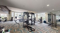 Located at 700 SW 78th Ave in Plantation, FL, this well-appointed fitness center offers a generously sized, single-level workout space designed to accommodate a wide range of exercise needs. The facility is defined by its soaring double-height ceilings in the cardio zone, which create an open, airy atmosphere, complemented by expansive floor-to-ceiling windows that flood the interior with natural light and frame lush outdoor views of tropical palms and landscaping. A striking large-scale mural accent wall adds a dynamic visual element to the space.

The fitness floor is covered in a stylish patterned carpet tile in neutral tones with teal accents, lending a modern and cohesive aesthetic throughout. Equipment is comprehensive and well-distributed, including treadmills, ellipticals, a stair climber, cable machines, a multi-station functional trainer, adjustable benches, a full dumbbell rack, barbells, and battle ropes, catering to both strength training and cardiovascular workouts. A dedicated stretching and functional training area features stability balls, step platforms, and exercise mats.

Large double doors provide direct exterior access, enhancing ventilation and connectivity to the outdoor amenity areas. Recessed lighting and wall-mounted TVs further elevate the facility's functionality and comfort, making this a thoroughly equipped and inviting fitness destination.