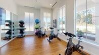 Located at 3030 Barrow Rd. in Charlotte, NC, this well-appointed fitness studio offers a bright and motivating environment for a full range of workouts. The single-floor space is designed with functionality and comfort in mind, featuring warm hardwood-style flooring throughout that adds a polished, inviting feel to the room.

Natural light pours in through multiple large windows, offering views of the surrounding greenery and creating an open, energizing atmosphere. High ceilings amplify the sense of space, while recessed lighting and a ceiling fan ensure comfort during intense workouts.

The studio is equipped with a row of Spinning bikes, ideal for cardio and cycling classes, as well as an air resistance bike for varied training options. A full wall of floor-to-ceiling mirrors enhances the sense of space and allows for form-checking during exercise. A large mounted flat-screen television is positioned on the mirror wall, making it easy to follow along with virtual fitness classes or training programs.

A dedicated yoga and stretching zone is clearly marked with signage, and colorful dumbbell racks provide a range of free weight options for strength training. Exercise balls are neatly organized on a storage rack, and a camera tripod setup suggests the space supports live or recorded fitness sessions. Clean white walls and crisp crown molding tie the design together with a modern yet classic aesthetic.