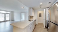 This single-floor space showcases a thoughtfully designed open-concept layout that seamlessly connects the kitchen and living area. The kitchen is appointed with crisp white cabinetry, light-toned quartz countertops, and a neutral tile backsplash in a subtle taupe hue that adds texture without overwhelming the clean aesthetic. A peninsula with generous counter space extends into the main living area, making it ideal for casual dining or entertaining. The stainless steel sink is equipped with a modern pull-down faucet, and a dishwasher is integrated neatly beneath the counter. Light wood-toned laminate flooring runs throughout, complementing the bright, white walls and contributing to the overall airy feel of the space. A track light fixture provides focused task lighting in the kitchen, while flush-mount ceiling lights illuminate the adjacent areas. A hallway off the kitchen leads to additional rooms, with an electrical panel access door visible along the corridor. The living area, visible beyond the kitchen, offers an open and versatile space filled with natural brightness. Overall, the unit presents a modern, neutral palette that is both functional and inviting.