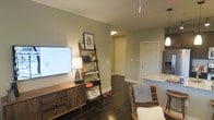 This single-floor space showcases a thoughtfully designed open-concept layout that connects the living area and kitchen in a cohesive, functional arrangement. The living area features rich, dark hardwood flooring that extends throughout, providing a warm contrast against the soft, neutral gray-toned walls. A wall-mounted flat-screen TV is positioned above a mid-century modern wooden media console, complemented by a decorative ladder shelf displaying framed artwork and accessories. A leather accent chair adds a touch of warmth to the seating area. The kitchen is partially visible to the right, featuring light granite countertops and modern cabinetry, with pendant lighting overhead contributing to the overall ambiance. A hallway visible in the background leads to additional areas of the unit, framed by clean white doors and trim. The space benefits from recessed and pendant lighting fixtures that ensure a well-lit interior. The overall design aesthetic is contemporary and cohesive, combining practical furnishings with tasteful décor elements that create a comfortable, inviting atmosphere throughout the open living space.