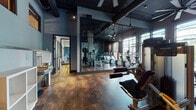Located at 100 Union St in Elizabeth, NJ, this fitness center combines industrial-contemporary design with a comprehensive array of workout amenities across a single, well-appointed floor. The space features rich hardwood-style flooring throughout, dark-toned walls, and exposed ceiling elements that lend an urban, loft-like atmosphere. Large floor-to-ceiling windows along the perimeter flood the cardio zone with abundant natural light while offering street-level views, creating an energizing environment for treadmill and elliptical workouts. Full-length mirrors are strategically placed to visually expand the space and support proper form during training. A dedicated spinning studio accommodates multiple stationary bikes and features a wall-mounted flat-screen TV and ceiling fan, making it ideal for group or individual cycling sessions. The free weights area is equipped with a tiered dumbbell rack, a weight bench, and additional strength machines, accented by bold abstract artwork that adds a creative, gallery-like touch. A separate strength and functional training zone houses cable machines and multi-use equipment. Convenient amenities include a dual-spout water fountain station and open storage. Ceiling fans throughout ensure comfortable airflow during workouts. The overall design balances high-end aesthetics with practical fitness functionality, making this facility a standout amenity.