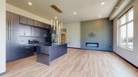 This single-floor residence offers a thoughtfully designed open-concept layout with high-end finishes and a cohesive modern aesthetic. The kitchen serves as the centerpiece, featuring dark cabinetry with gold hardware, a bold navy island with a dark quartz countertop, a stainless steel undermount sink with a gold pull-down faucet, and stainless steel appliances including a refrigerator and range. A striking cluster of gold-framed pendant lights cascades above the island, adding a dramatic focal point. The kitchen flows seamlessly into the living area, which is anchored by a textured accent wall and a sleek, built-in linear fireplace, ideal for gathering. Light wood-tone luxury vinyl plank flooring extends throughout the main living spaces, complementing the contemporary palette. The bedrooms are finished with plush carpeting and feature large windows that frame verdant outdoor views, filling each room with natural light. Soaring ceilings in the bedroom areas enhance the sense of spaciousness. A dedicated laundry room includes full-size washer and dryer units alongside a custom dark built-in cubby and bench system with gold hooks, functioning as a practical mudroom. A full bathroom with wood-tone vanity, walk-in shower, and wood-look flooring rounds out the space with clean, functional design.