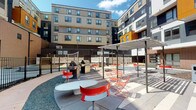 This outdoor courtyard amenity space is nestled within a contemporary multi-story residential building complex, offering a well-appointed communal environment designed for social gathering and outdoor living. The courtyard is framed on multiple sides by the building's modern façade, which features a striking combination of neutral panels, dark cladding, and bold orange accent sections, creating a visually dynamic architectural backdrop.

At the heart of the space, a dedicated grilling station is equipped with multiple full-sized gas grills, providing ample capacity for group cooking and outdoor entertaining. Adjacent to the grills, a sleek pergola structure offers shaded seating beneath an open lattice canopy, furnished with vibrant red folding chairs and contemporary round tables — adding a lively pop of color to the overall aesthetic.

The ground level is thoughtfully finished with a mix of paved walkways, gravel ground cover, and defined seating zones that encourage both intimate gatherings and larger social events. Perimeter fencing adds a sense of enclosure and privacy to the courtyard without compromising its open, airy atmosphere. Under open skies, the space benefits from abundant natural light throughout the day, making it an inviting retreat for residents seeking an active and connected outdoor experience.