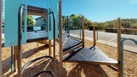 This outdoor recreational space centers around a well-equipped playground structure designed for active play. The play structure features multiple components, including a curved slide, a textured climbing column with footholds, a ramped access bridge with non-slip perforated metal grating, and teal-colored panel enclosures with secure handrails. The entire playground area is surfaced with a generous layer of wood chip mulch, providing a safe and impact-absorbing ground cover throughout.

The surrounding grounds are thoughtfully landscaped, featuring neatly maintained green lawn areas bordered by concrete pathways. A fenced patio area adjacent to a single-story building with a distinctive red door offers outdoor seating beneath a red umbrella, with chairs and tables arranged for casual gatherings. Potted plants and a wooden bench add a welcoming touch near the building entrance.

A covered pavilion with an open-air design is situated nearby, offering a shaded gathering space. The perimeter is lined with mature trees and well-trimmed hedges, providing natural screening and shade. Under clear blue skies, the entire space benefits from abundant natural light, creating a bright and inviting outdoor environment suitable for both recreation and relaxation.