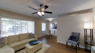 This single-floor space showcases an open-concept layout that seamlessly connects the living and dining areas, creating a cohesive and functional flow. The living area features warm hardwood-style flooring that extends throughout, anchored by a plush area rug and complemented by neutral-toned walls that lend a clean, inviting atmosphere. A ceiling fan with an integrated light fixture serves as a practical centerpiece overhead, while a textured popcorn ceiling adds subtle depth to the space.

Natural light filters in generously through multiple windows, including a large vertical-blind-covered window that frames an outdoor view, brightening the interior throughout the day. The dining area, partially open to the living space, is defined by its own overhead light fixture and accommodates a glass-top dining table with upholstered chairs. Decorative wall art adds a touch of personality to the dining nook. Clean white baseboards frame the flooring throughout, adding a crisp finish to the overall design. The space offers a comfortable, functional layout well-suited for everyday living and entertaining.