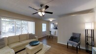 This single-floor space showcases an open-concept layout that seamlessly connects the living and dining areas, creating a cohesive and functional flow. The living area features warm hardwood-style flooring that extends throughout, anchored by a plush area rug and complemented by neutral-toned walls that lend a clean, inviting atmosphere. A ceiling fan with an integrated light fixture serves as a practical centerpiece overhead, while a textured popcorn ceiling adds subtle depth to the space.

Natural light filters in generously through multiple windows, including a large vertical-blind-covered window that frames an outdoor view, brightening the interior throughout the day. The dining area, partially open to the living space, is defined by its own overhead light fixture and accommodates a glass-top dining table with upholstered chairs. Decorative wall art adds a touch of personality to the dining nook. Clean white baseboards frame the flooring throughout, adding a crisp finish to the overall design. The space offers a comfortable, functional layout well-suited for everyday living and entertaining.
