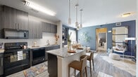 This single-floor residence showcases a contemporary design with a cohesive color palette of cool blues, crisp whites, and warm wood tones carried throughout. The open-concept kitchen and living area form the heart of the home, where the kitchen stands out with dark, textured flat-front cabinetry reaching toward the ceiling, granite countertops, a mosaic tile backsplash, and a full suite of black appliances. A central island with a granite surface and bar seating serves as a natural gathering point, illuminated by a trio of modern cylindrical pendant lights that add visual drama overhead. Light wood-style flooring runs continuously through the main living areas, enhancing the sense of openness.

The living room is characterized by deep blue-grey accent walls, a ceiling fan, and a bright window that invites natural light. A built-in shelving unit adds a functional and decorative focal point alongside the casual seating arrangement.

The bedroom continues the blue accent wall theme, featuring large windows dressed with layered curtains, plush carpeting, and a ceiling fan. The bathroom is finished with the same granite countertop and dark cabinetry as the kitchen, paired with wood-style flooring and a tiled shower, maintaining a consistent, polished aesthetic throughout the residence.