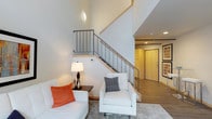 This thoughtfully designed two-story residence combines modern aesthetics with functional living across two levels connected by a sleek staircase. The living area makes an immediate impression with soaring ceilings that amplify the sense of openness, complemented by clean white walls and warm wood-toned accents along the staircase railings. The staircase itself features metal balusters with light wood detailing, serving as both a functional and architectural focal point. Light wood flooring flows throughout, lending warmth and continuity to the space, while recessed lighting and a stylish wall-mounted lamp provide layered illumination. The entryway is bright and welcoming, finished with a natural wood door that adds a warm contrast to the contemporary palette.

The building's communal laundry facility is a standout amenity, housed in a space with striking industrial-chic character. Soaring ceilings showcase exposed wooden roof trusses, visible ductwork, and original brick accent walls, evoking the charm of a thoughtfully converted historic structure. A row of commercial-grade stainless steel washers and dryers lines one wall, while large windows flood the room with natural light. A cozy seating area with upholstered chairs and a round side table offers a comfortable place to wait, rounding out this well-equipped shared space.