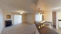 This single-floor space features an open-concept layout that seamlessly connects the kitchen and living areas. The living room is finished with plush carpeting and is anchored by a ceiling fan, contributing to a comfortable and functional atmosphere. Sliding glass doors open to a private balcony, allowing natural light to fill the space while offering a direct connection to the outdoors. The kitchen area transitions from wood-style flooring and includes pendant lighting overhead, with a dark-toned countertop visible at the edge of the frame. Neutral beige walls run throughout, complemented by a textured popcorn ceiling that adds dimension overhead. HVAC vents are positioned throughout for efficient climate control. A hallway visible from the main living area leads to additional rooms, including what appears to be a bathroom with natural light. The overall palette of warm neutrals and the open flow between spaces create a cohesive, practical environment suited for everyday living. The combination of carpeted and hard-surface flooring effectively delineates functional zones within the layout.
