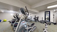 This fitness center in Walnut Creek, CA (94596) offers a thorough and well-organized workout environment spread across a single floor. The space is outfitted with patterned, cushioned carpeting throughout, providing comfort underfoot during exercise sessions. Natural light filters in through a series of horizontal windows along the upper walls, complemented by recessed and flush-mounted ceiling fixtures that keep the space well-lit at all times.

The cardio section is prominently featured, housing multiple Star Trac elliptical trainers and treadmills, as well as a stationary spin bike, allowing users a variety of aerobic options. Wall-mounted flat-screen televisions are positioned throughout the space, offering entertainment during workouts. A dedicated strength-training area includes a multi-station cable machine, a leg press, and additional resistance equipment. The free weights section features a full dumbbell rack, kettlebells, barbells, and weight plates neatly organized along the wall. Exercise balls in multiple sizes round out the functional fitness offerings.

Full-length mirrors are mounted along one wall, enhancing the sense of space and supporting proper form during exercise. The neutral white walls and textured ceilings create a clean, focused atmosphere suited for an effective and comfortable fitness experience.