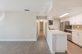 This single-floor residence offers a thoughtfully designed open-concept layout that seamlessly connects the main living areas. The entryway welcomes you with warm-toned hardwood-style flooring that extends throughout the main living and kitchen spaces, creating a cohesive and inviting atmosphere.

The kitchen is a functional highlight of the home, featuring white shaker cabinetry, a clean white countertop peninsula that doubles as a breakfast bar, and a full suite of stainless steel appliances including a gas range, over-the-range microwave, and refrigerator. A decorative tile backsplash adds a subtle touch of texture and style to the cooking area.

The open living area flows naturally from the kitchen, offering generous space bathed in neutral tones with warm gray walls and crisp white trim. A front-facing window allows natural light to filter in, enhancing the bright and airy feel of the space.

The bedroom is carpeted for comfort and features a sliding glass door fitted with vertical blinds, offering access to an outdoor area and welcoming an abundance of natural light.

The bathroom is well-appointed with a white vanity, a framed mirror, bright overhead lighting, and a full tub-shower combination, all complemented by hardwood-style flooring that ties the room together cleanly.