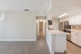 This single-floor residence offers a thoughtfully designed open-concept layout that seamlessly connects the main living areas. The entryway welcomes you with warm-toned hardwood-style flooring that extends throughout the main living and kitchen spaces, creating a cohesive and inviting atmosphere.

The kitchen is a functional highlight of the home, featuring white shaker cabinetry, a clean white countertop peninsula that doubles as a breakfast bar, and a full suite of stainless steel appliances including a gas range, over-the-range microwave, and refrigerator. A decorative tile backsplash adds a subtle touch of texture and style to the cooking area.

The open living area flows naturally from the kitchen, offering generous space bathed in neutral tones with warm gray walls and crisp white trim. A front-facing window allows natural light to filter in, enhancing the bright and airy feel of the space.

The bedroom is carpeted for comfort and features a sliding glass door fitted with vertical blinds, offering access to an outdoor area and welcoming an abundance of natural light.

The bathroom is well-appointed with a white vanity, a framed mirror, bright overhead lighting, and a full tub-shower combination, all complemented by hardwood-style flooring that ties the room together cleanly.