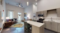 This thoughtfully designed single-floor residence immediately impresses with its soaring ceilings that lend an airy, expansive quality to the open-concept living and kitchen area. The kitchen is well-appointed with stainless steel appliances, including a range, microwave, and refrigerator, complemented by sleek wood-grain cabinetry, white quartz countertops, and a central island with bar seating — ideal for casual dining or entertaining. The living room flows naturally from the kitchen and is anchored by a large decorative area rug, a ceiling fan, and a glass-paned exterior door that opens to an outdoor balcony, allowing ample natural light to filter in. The space includes two bedrooms, each carpeted and fitted with windows dressed in patterned curtains, offering a comfortable and private retreat. The bathrooms feature modern tile flooring in a herringbone-inspired layout, wood-grain vanity cabinetry with white countertops, and generous storage including a tall linen cabinet. A walk-in closet is accessible from the primary bathroom. Throughout the residence, a consistent palette of neutral tones, contemporary fixtures, and clean lines creates a cohesive and polished aesthetic suited to modern living.