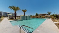Located at 17120 North 53rd Avenue in Glendale, Arizona, this modern two-story residential community is designed around an expansive, resort-style outdoor amenity space that serves as its centerpiece. The architecture features a contemporary multi-story building with clean lines, neutral stucco exteriors, private balconies, and large windows that overlook the courtyard below.

The outdoor area is anchored by a generously sized swimming pool with striking tile water-wall features, providing both visual appeal and a cooling ambiance suited to the Arizona climate. Mature palm trees are strategically positioned throughout the pool deck, offering shade and a resort-like atmosphere under the abundant Sonoran sunshine. Multiple private cabanas with retractable canopy covers and striped curtains line the pool perimeter, offering shaded lounging options.

A dedicated outdoor kitchen and bar area, covered by a modern pergola-style canopy, provides seating and built-in grilling facilities for outdoor dining and entertaining. Adjacent to the pool, a well-maintained turf lawn is furnished with contemporary lounge seating and large umbrellas, creating a comfortable gathering space. Decorative desert landscaping with native plants and gravel grounds the overall design aesthetic. The community's layout, as viewed from above, reveals a thoughtfully planned courtyard configuration that promotes an open, connected outdoor living environment.