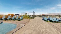 Located at 55 N Park Ave in Tucson, Arizona, this rooftop amenity deck offers a striking outdoor retreat above the Tucson skyline. The centerpiece of the space is a generously sized resort-style swimming pool with crystal-clear water, complemented by an adjacent spa that together create a cohesive aquatic experience. The pool deck is finished with durable, light-toned paver tiles that extend throughout the space, providing a clean and modern aesthetic underfoot. Rows of sleek lounge chairs line the perimeter of the pool, offering ample opportunity for sunbathing and relaxation in Tucson's abundant sunshine. Planters featuring desert-appropriate vegetation, including cacti and ornamental grasses, add natural texture and a nod to the local landscape. String lights are strung overhead in a designated social area, creating an inviting atmosphere for evening gatherings. Glass railings along the deck's edges preserve unobstructed panoramic views of the Santa Catalina Mountains to the north and the broader Tucson cityscape. The surrounding modern multi-story architecture frames the space while reinforcing the urban setting. The open-air design maximizes exposure to the Southwest's expansive blue skies, making this rooftop deck a standout amenity that blends leisure, design, and natural scenery seamlessly.
