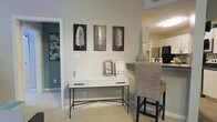 This single-floor residence offers a thoughtfully designed layout that balances comfort and modern style. The entryway sets an inviting tone with neutral-toned walls, light carpeting, and curated wall art featuring decorative leaf panels that add a natural, artistic touch. A sleek white console table with metal legs anchors the space, providing both functionality and visual interest.

The open-concept flow connects the entry area seamlessly to the adjacent kitchen, which is visible in the background and features standard cabinetry, modern appliances, and a raised bar counter. A woven rattan bar stool and a tall decorative vase add texture and warmth to the transitional space between rooms.

A hallway visible from the main area leads to additional rooms, hinting at a well-organized floor plan. The walls throughout are finished in soft, neutral tones that complement the light flooring, creating a cohesive and airy ambiance. Flush-mounted ceiling lighting provides consistent illumination across the space.

Overall, this residence presents a clean, contemporary aesthetic with carefully chosen decorative elements that enhance its welcoming character. The layout is practical and accessible, making efficient use of the available space while maintaining a polished, livable atmosphere throughout.