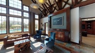 Located at 12150 Washington Center Pkwy in Thornton, Colorado, this single-story community clubhouse delivers an impressive blend of rustic elegance and modern amenity. The main gathering area commands immediate attention with its dramatic vaulted ceiling showcasing exposed dark wood truss work, complemented by large pendant light fixtures. Floor-to-ceiling multi-pane windows flood the space with natural light and offer direct views of the outdoor pool area, creating a seamless indoor-outdoor connection. A stately stacked-stone fireplace with a solid wood mantle anchors one end of the lounge, which is furnished with tufted leather sofas and rich blue velvet wingback chairs atop a decorative area rug and mixed slate tile flooring. A built-in dark wood media wall with cabinetry provides a focal display area. The well-appointed kitchen features warm golden granite countertops, rich mahogany cabinetry, stainless steel appliances including a large refrigerator and double wall oven, and a gas cooktop. A separate game room offers a full-size pool table, a built-in entertainment wall with cabinetry, a flat-screen TV, and a bar-height counter with seating — ideal for social gatherings. Throughout, the design maintains a cohesive, upscale lodge-inspired aesthetic.