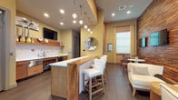 This single-floor space functions as a well-appointed lounge or clubroom, blending contemporary design with warm, inviting materials. The centerpiece is a substantial bar-height island with a live-edge wood base and bright white countertop, accompanied by upholstered white bar stools with nailhead detailing. A cascade of globe pendant lights suspended from a stepped ceiling soffit illuminates the island, adding a striking visual focal point.

The kitchenette area along one wall features warm wood cabinetry, a sink, stainless steel appliances, open floating shelves with decorative accents, and a wall-mounted television. Dark hardwood-style flooring runs throughout, grounding the warm yellow-toned walls. A textured wood plank accent wall anchors a dedicated lounge corner, where two flat-screen televisions are mounted above round white tables flanked by studded dark upholstered chairs.

Natural light filters in through large windows dressed with neutral blinds and sheer curtains, while recessed lighting ensures consistent illumination throughout. A decorative mirrored disc wall sculpture adds an artistic touch. Double glass-paned entry doors connect to an adjacent corridor, reinforcing the communal nature of the space. The overall ambiance is modern, warm, and social.