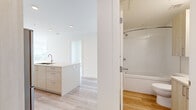 This well-designed 3-bedroom, 2-bathroom single-floor residence offers a clean, contemporary aesthetic throughout. The open-concept living area is bathed in natural light, thanks to floor-to-ceiling windows that frame impressive panoramic views of the surrounding cityscape and skyline. Light hardwood flooring runs consistently throughout the space, complementing the crisp white walls and creating a bright, airy ambiance.

The kitchen features a functional island with an undermount sink, white cabinetry, and recessed lighting, flowing seamlessly into the living area for an integrated feel. Each of the three bedrooms is finished with the same light hardwood flooring and neutral walls, with windows that bring in natural light and outdoor views of the neighborhood. The primary bedroom offers a comfortable footprint suitable for everyday living.

The two bathrooms are finished in clean white tiling. One features a soaking tub with a tiled surround and a sliding glass shower enclosure, while the other includes a glass-enclosed stand-alone shower with polished chrome fixtures. A connecting hallway ties the layout together efficiently. Overall, the space delivers a minimalist, modern living experience with an emphasis on natural light, clean finishes, and functional design.
