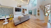 Located at 109 W Fortune St in the heart of downtown Tampa, this contemporary amenity space blends modern design with vibrant, welcoming interiors. The main lounge area commands attention with its soaring double-height ceilings, floor-to-ceiling windows, and an abundance of natural light that floods the warm hardwood floors below. Oversized globe pendant lights hang dramatically overhead, complementing a large wall-mounted TV framed by built-in wood display niches showcasing decorative vases and accents.

A standout lounge nook features a striking textured mosaic accent wall in a shimmering penny-tile finish, backed by a curved teal sectional sofa and intimate bistro seating with pops of yellow, creating a cozy yet lively atmosphere. A generous communal dining island with a light wood surface and wire café-style chairs provides a versatile gathering and co-working area, flanked by tall sculptural branch arrangements in elegant white vases.

The kitchenette is outfitted with wood-toned countertops, white cabinetry with gold hardware, a built-in microwave, refrigerator drawers, and a sink — all set against a bold geometric patterned tile backsplash. Glass doors open to the outdoors, reinforcing the bright, airy character of this thoughtfully designed community space.