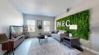This thoughtfully designed single-floor unit at Parc offers a complete living experience within a compact footprint, comprising a living room, kitchen, and bathroom. The living room serves as the centerpiece of the space, immediately drawing the eye to a dramatic full-wall moss installation bearing the "PARC" lettering — a bold, nature-inspired accent that adds life and texture to the neutral gray walls. Light wood-style flooring runs seamlessly throughout, enhancing the sense of openness. Multiple windows invite natural light and views of surrounding greenery, contributing to a bright and airy ambiance. The galley-style kitchen runs along one wall and is efficiently appointed with dark cabinetry, a stainless steel refrigerator, stove, and microwave, complemented by light countertops and a single-basin sink. A console table and large abstract artwork create a stylish transitional zone between the kitchen and the entry door. The bathroom is clean and simply finished, featuring a shower-tub combination with a neutral striped curtain. Throughout the unit, the design balances modern aesthetics with functional use of space, creating an inviting and cohesive living environment.