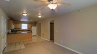 This single-floor space features an open-concept design that seamlessly connects the living area and kitchen, creating a cohesive and functional layout. The living area is finished with wall-to-wall carpet in a warm neutral tone, contributing to a comfortable and inviting atmosphere. A ceiling fan with an integrated light fixture is centrally positioned overhead, providing both air circulation and ambient lighting. Flush-mount light fixtures are also installed throughout, ensuring consistent illumination across the space.

The kitchen is defined by light wood cabinetry, a countertop with a built-in sink, and tile flooring that contrasts neatly with the carpeted living area. A full-size refrigerator and additional appliances are included. A window above the sink allows for natural light to filter into the kitchen area.

Adjacent to the living space, a doorway leads to a walk-in closet fitted with a clothing rod, offering practical storage. The walls throughout are painted in a soft, warm beige, and clean white baseboards provide a crisp finish. A floor-level vent is also present, indicating a central HVAC system for year-round climate control.
