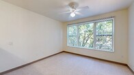 This efficiently designed single-floor studio offers a well-organized layout that maximizes every square foot of its compact footprint. The main living area features soft, neutral-toned carpeting underfoot and a ceiling fan overhead, creating a comfortable and airy atmosphere. A large picture window fitted with vertical blinds fills the space with natural light and frames a pleasant view of surrounding tree canopy, bringing a sense of the outdoors inside.

The kitchen is laid out in a practical galley style, featuring light wood cabinetry on both upper and lower walls, a white electric range, a full-size refrigerator, a stainless steel sink with ample counter space, and wood-look flooring that adds warmth to the room. The kitchen is also accessible from a counter-height breakfast bar area, offering an open connection to the main living space.

A dedicated hallway leads to built-in closet storage with wire shelving and hanging rods, providing organized space for personal belongings. The bathroom is clean and functional, appointed with a wood-toned vanity, a mirror, a toilet, and wood-look tile flooring consistent with the rest of the unit's finishes. Warm neutral tones throughout the space lend a cohesive and understated aesthetic, making this unit an practical and comfortable living solution.