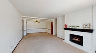 This single-floor residence offers a practical and comfortable layout designed for easy living. The expansive living area is the centerpiece of the home, featuring plush neutral carpeting, a striking white brick fireplace with a dark mantel, and an open flow into the dining area, which is equipped with a ceiling fan and exposed ceiling beams. Large vertical blinds and windows allow natural light to fill the space throughout the day.

The kitchen is efficiently designed with warm wood cabinetry, light countertops, a double basin sink, and black appliances including a range and dishwasher. A stacked washer and dryer unit is conveniently integrated within the kitchen area, adding everyday functionality.

The bedrooms are generously sized, each finished with soft carpeting, baseboard heating, and windows with blinds that provide natural light and privacy. The rooms offer ample wall space and a clean, neutral palette suitable for a variety of uses.

Two full bathrooms serve the residence. One features a tub-shower combination with a curtain, tile flooring, and a warm wood vanity with light countertops. The second bathroom offers a similar vanity setup and includes a separate tub area. Both bathrooms are finished with tile flooring and provide functional, well-organized layouts. Overall, the home presents a clean and move-in ready environment.